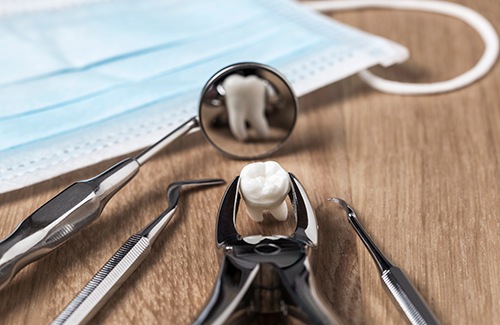 Extracted tooth grasped by forceps reflected in dental mirror on desk next to face mask
