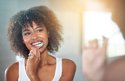 Woman in white tank top with curly brown hair brushing her teeth in front of a mirror