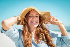 Woman with red hair wearing straw hat at the beach smiling with eyes closed